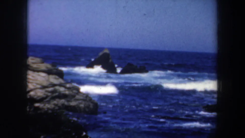 1957 San Francisco 8mm frame showing blue ocean surf breaking around dark offshore rocks and pale coastal stone.