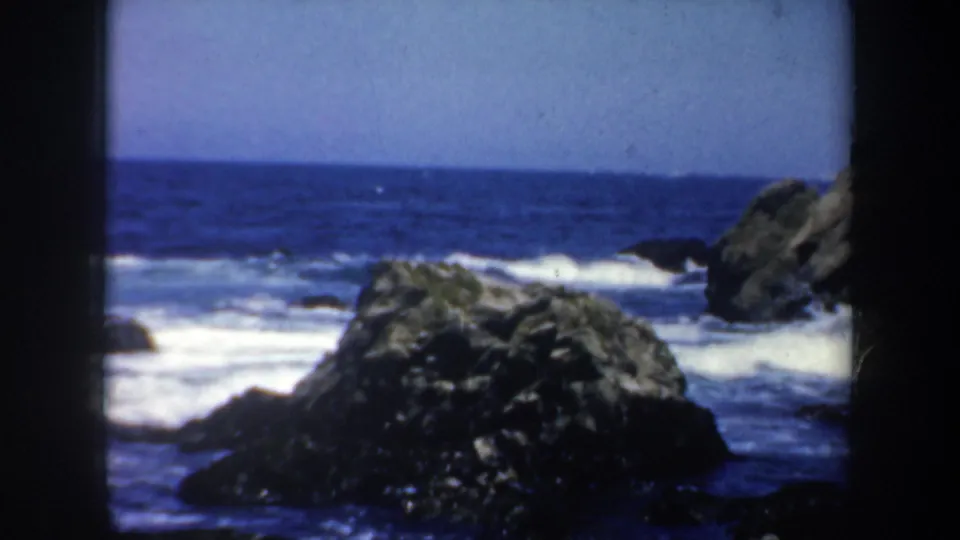 1957 San Francisco coastal frame showing a large foreground rock, white surf, and blue ocean under a clear sky.
