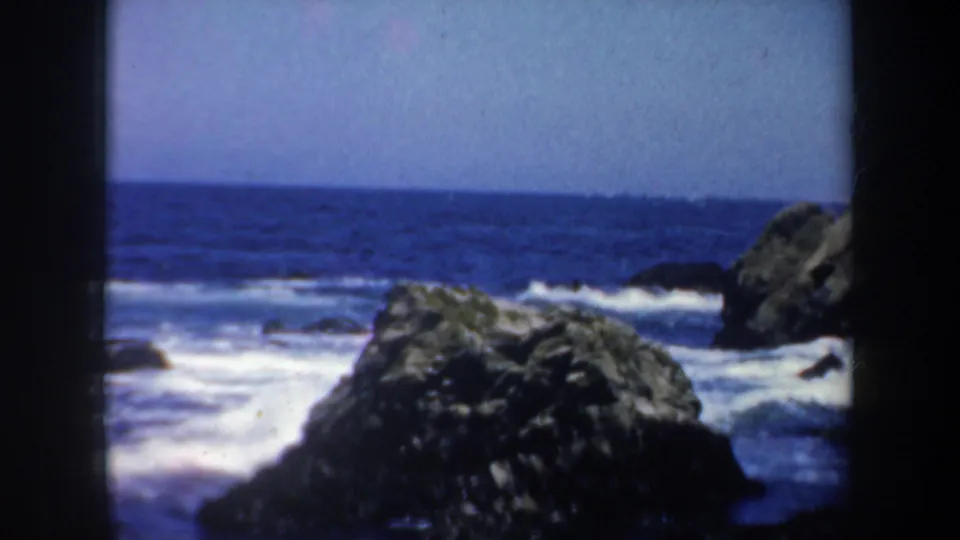 1957 San Francisco coastal home-movie frame showing dark boulders, blue water, white surf, and visible 8mm frame edges.
