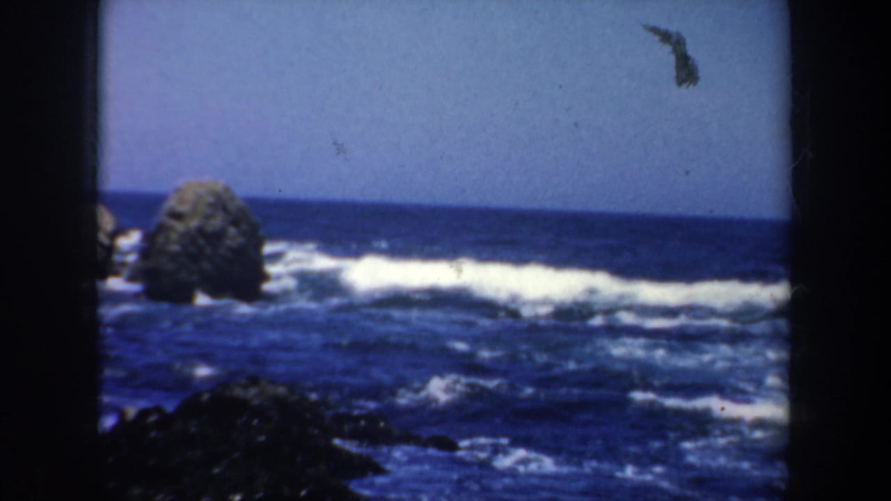 1957 San Francisco coastline frame with offshore boulders, blue sea, white surf, and dark 8mm gate edges.