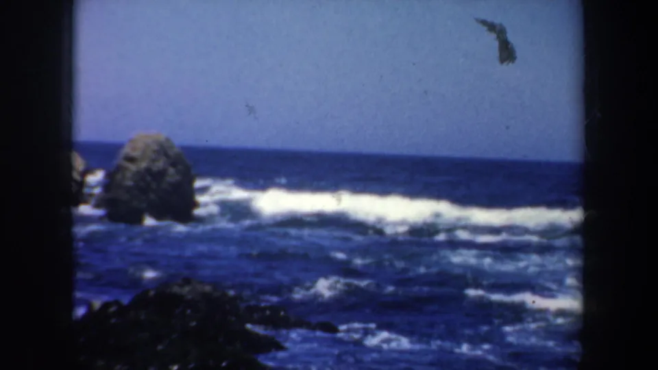 1957 San Francisco coastline frame with offshore boulders, blue sea, white surf, and dark 8mm gate edges.