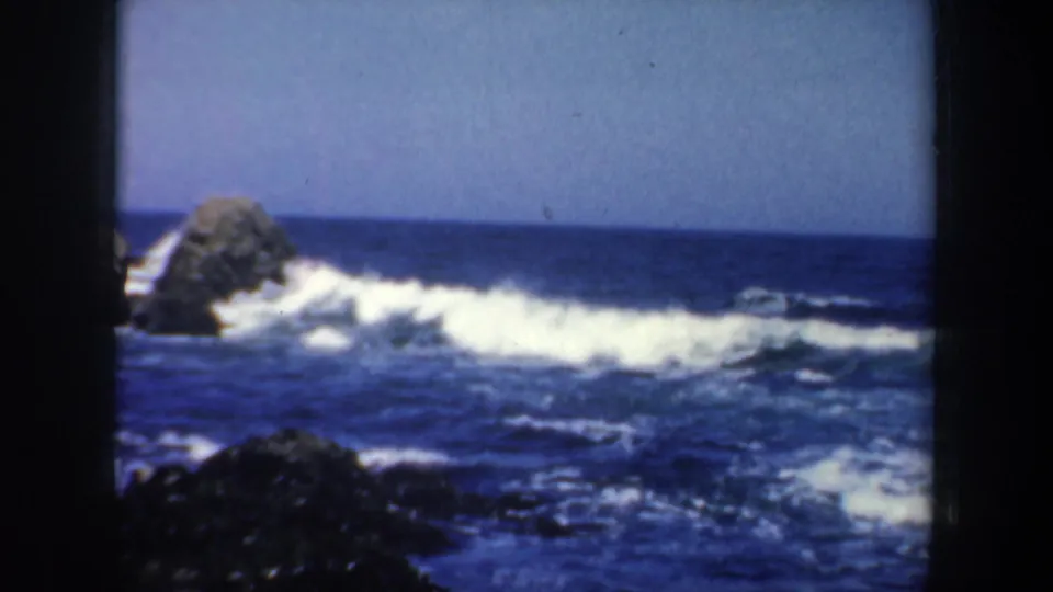 1957 San Francisco coastal frame showing white waves, dark foreground rocks, blue water, and a large offshore boulder.