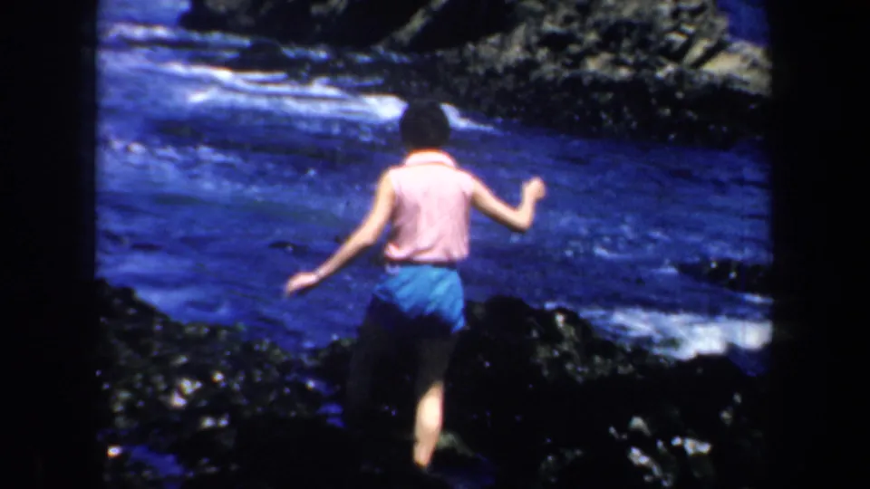 1957 San Francisco home-movie frame showing a person from behind walking across dark coastal rocks near blue surf.