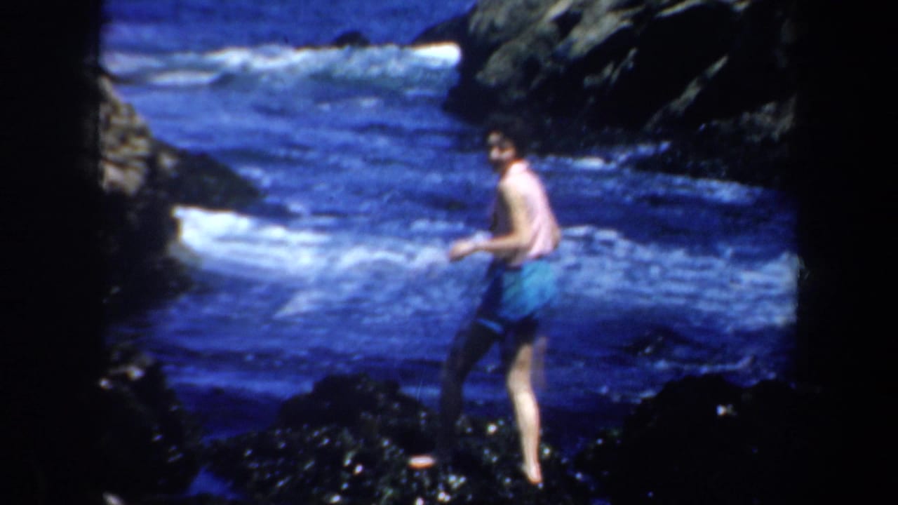 1957 San Francisco home-movie frame showing an adult in pink and blue swimwear balancing on dark shoreline rocks.