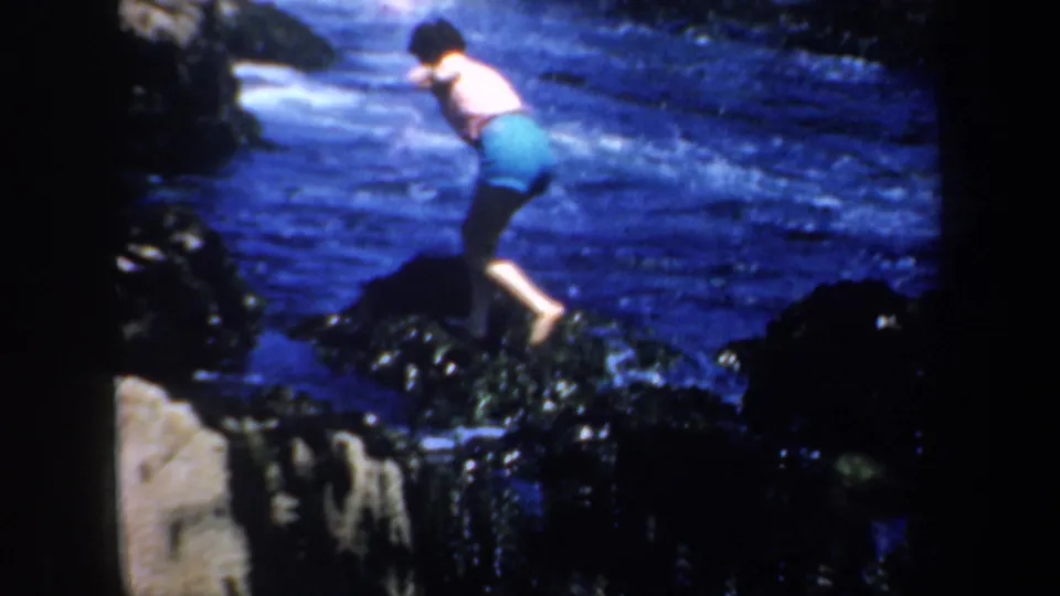 1950s coastal frame of an adult swimmer crossing dark, slippery rocks with blue surf behind.