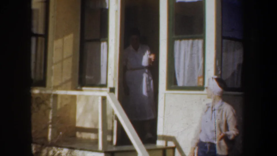 A woman in a light dress stands partly inside the doorway as another adult woman in a headscarf passes below the porch.