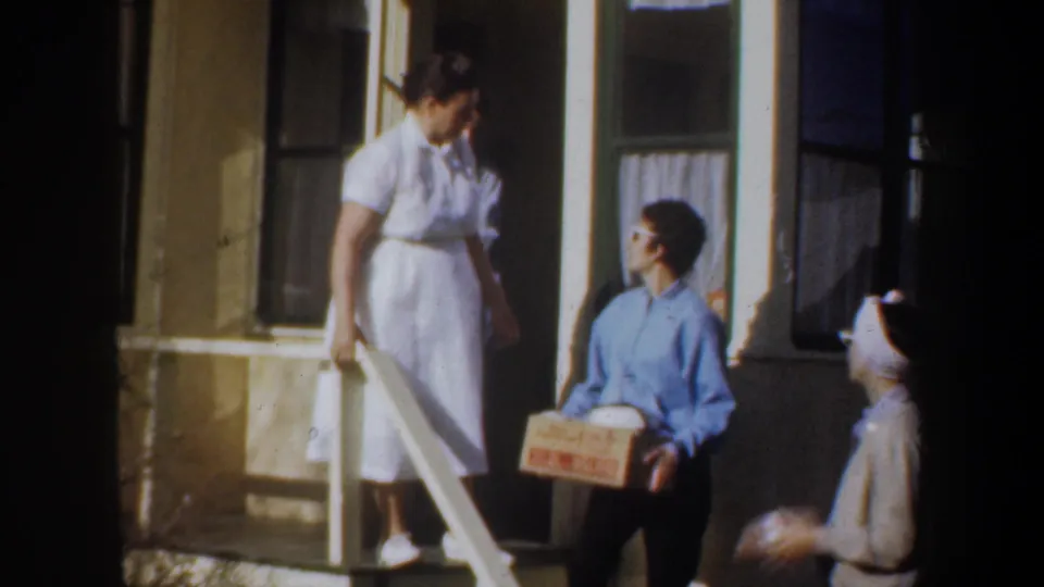 On the Portland porch, an adult woman in a white dress looks toward a seated woman holding a cardboard box near the steps.