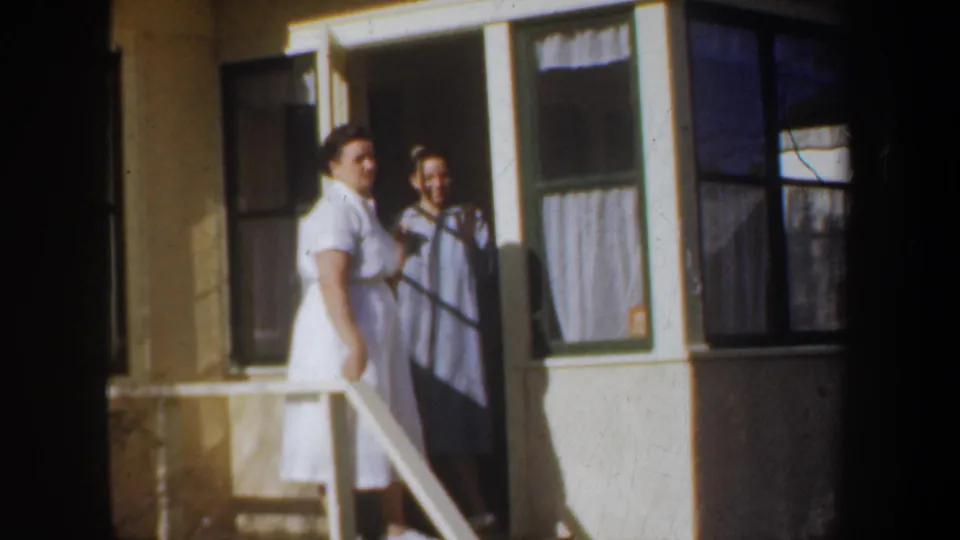 Two women on a Portland porch in 1957 appear to pause during a friendly goodbye, with house windows and a white railing behind them.