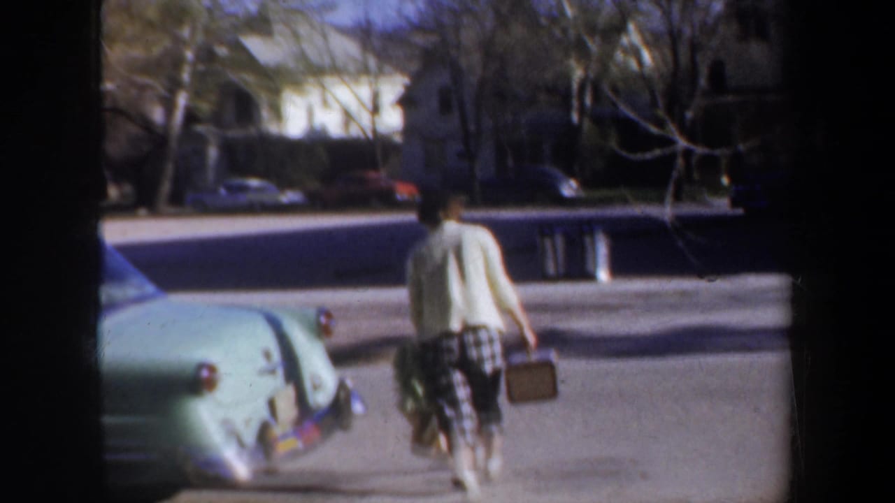 A woman walks away on a residential street carrying luggage near a pale green car.