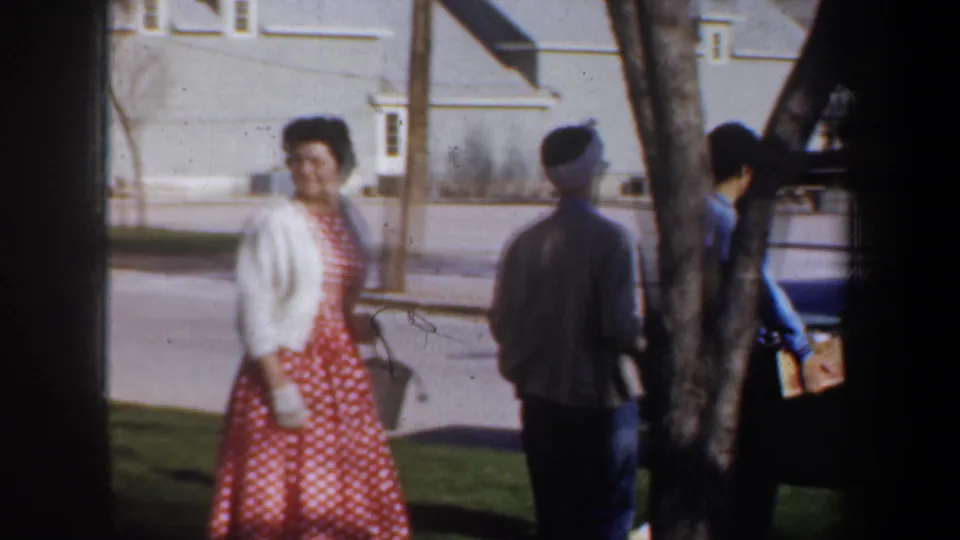 A woman in a red polka-dot dress and white cardigan walks with others in a sunny 1957 Portland residential setting.