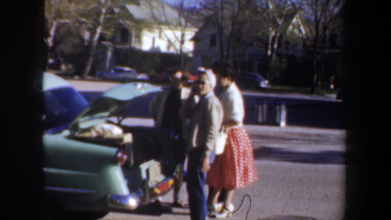 1957 Portland home-movie frame showing adults by an open-trunk light green car during apparent road-trip packing.