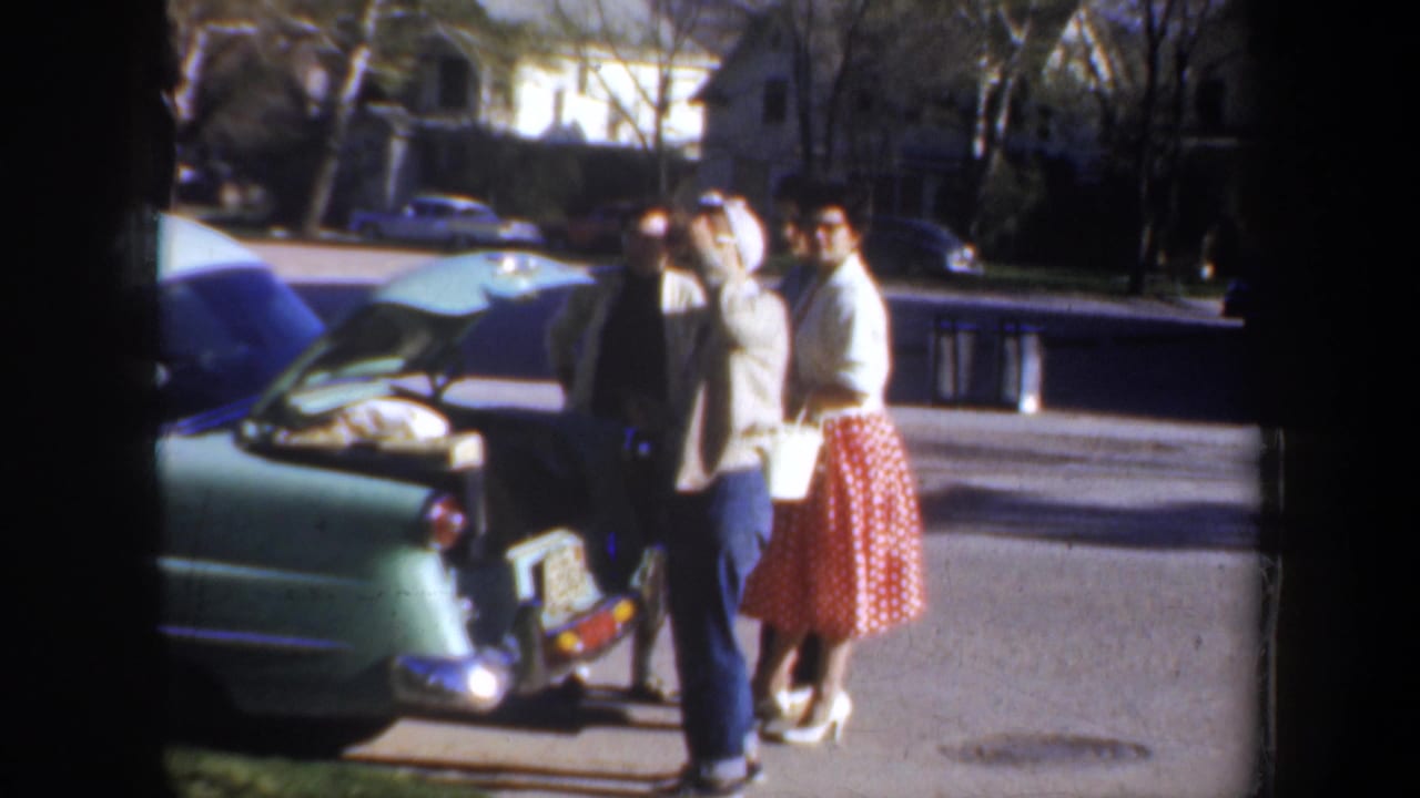 1957 Portland scene with adults beside a packed light green car, including a woman in a red polka-dot skirt and another adjusting a headscarf.