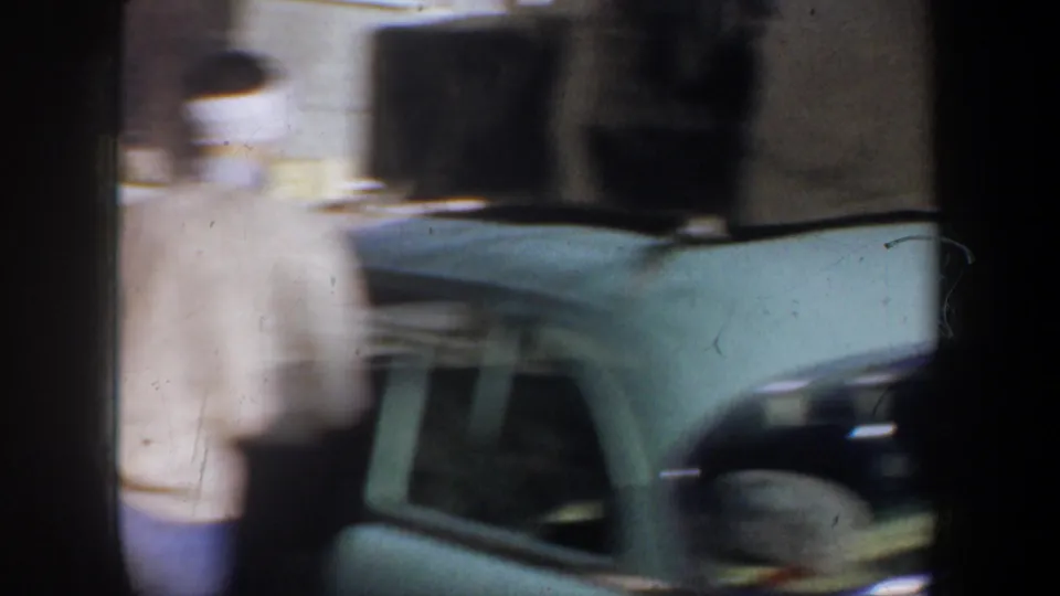 Blurred 8mm frame from the Portland road-trip scene, with a person moving past the light green car and luggage on top or near the vehicle.