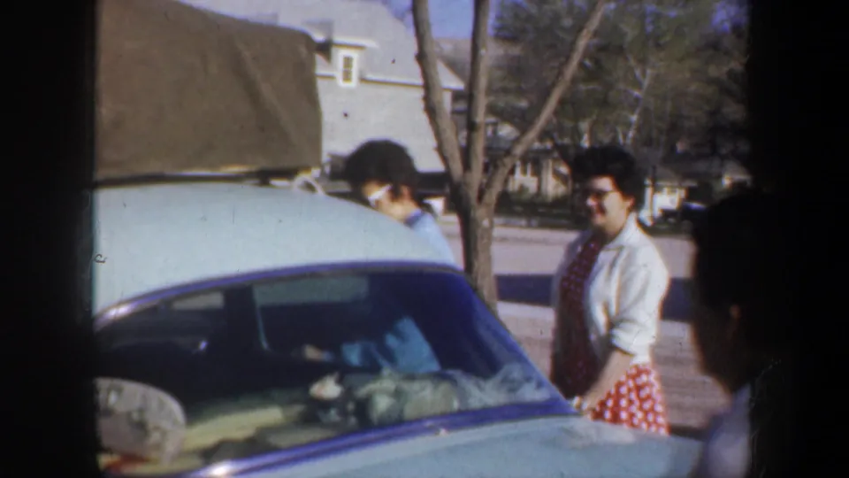 1957 Portland frame showing adults by the light green car, with packed luggage visible and a woman in a red polka-dot skirt at right.
