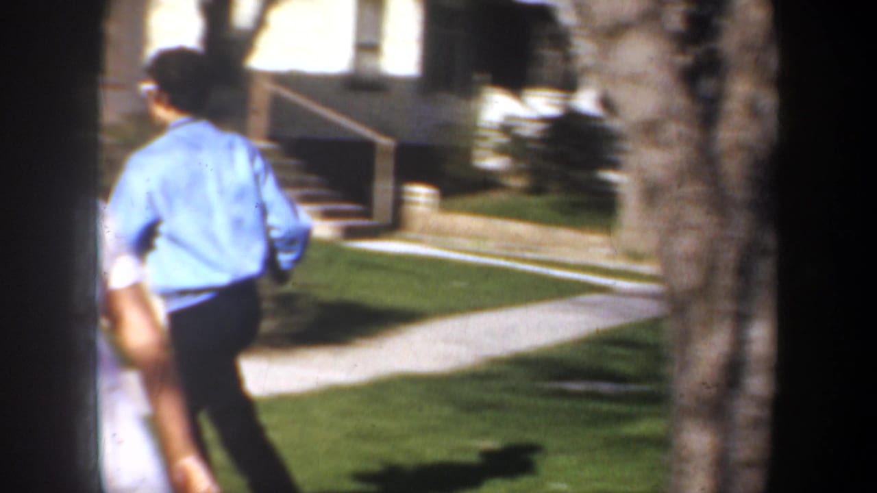 1957 Portland home-movie frame showing two adults walking through a suburban yard near trees and a concrete path.