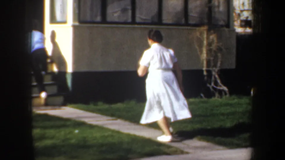 1957 Portland home-movie frame showing a woman in white walking behind a man near steps and a house exterior.