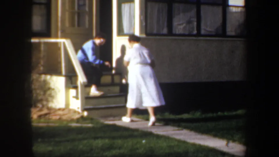 1957 Portland exterior frame with a man bending at the steps and a woman standing nearby on the yard path.