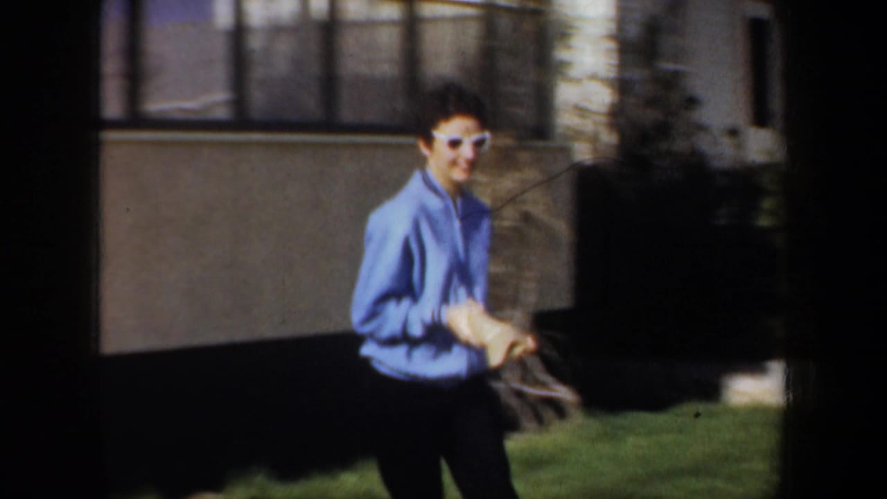 In Portland, Oregon home-movie footage from 1957, a woman in a blue sweater and white sunglasses moves across a front yard.