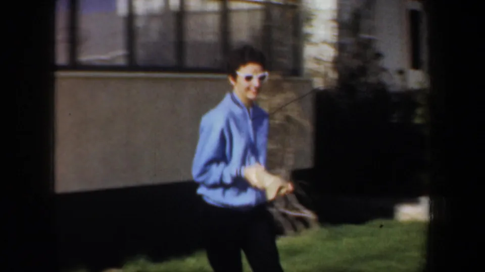 In Portland, Oregon home-movie footage from 1957, a woman in a blue sweater and white sunglasses moves across a front yard.