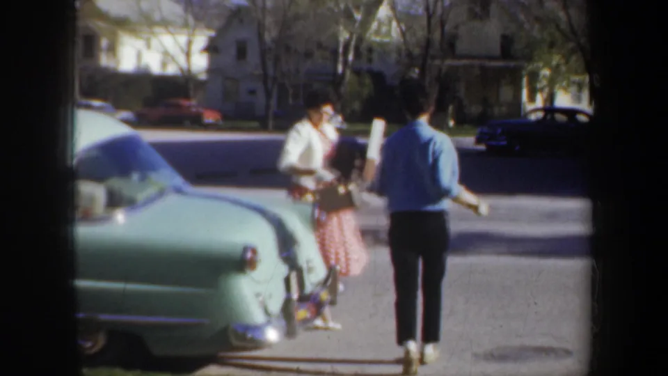 Two women approach each other beside a pale green car on a Portland residential street in 1957 home-movie footage.