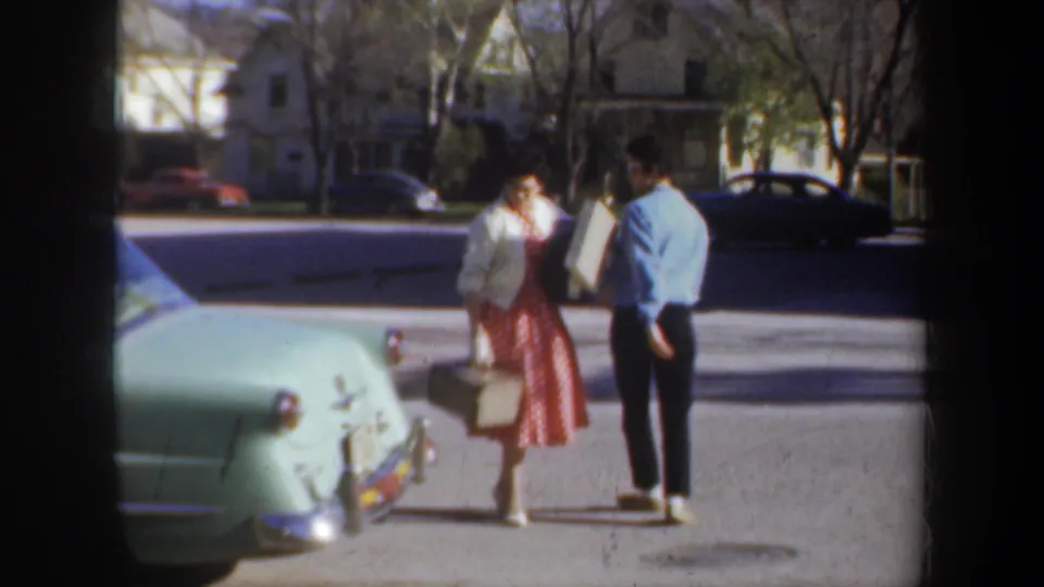 The two women pause near the rear of a pale green car, with houses and parked cars visible across the Portland street.