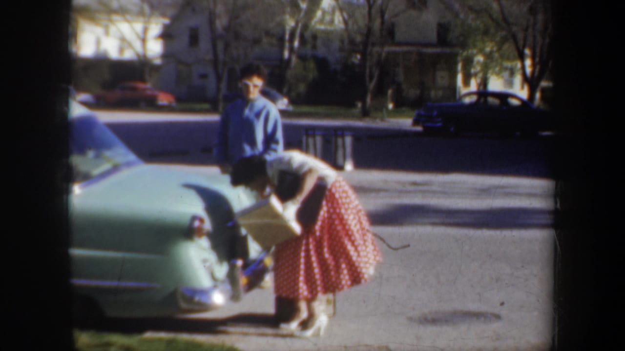 In metadata-supported 1957 Portland, a woman in a red polka-dot skirt leans toward an open car trunk as a man in a blue shirt watches nearby.