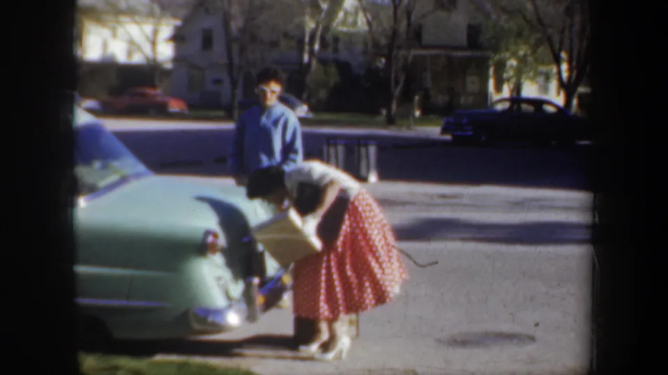 In metadata-supported 1957 Portland, a woman in a red polka-dot skirt leans toward an open car trunk as a man in a blue shirt watches nearby.