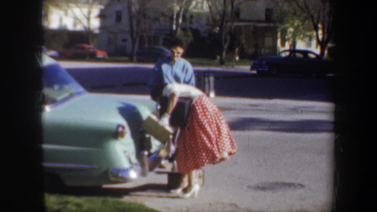 Woman leans over the car trunk with a box as a man stands behind her near the curb.