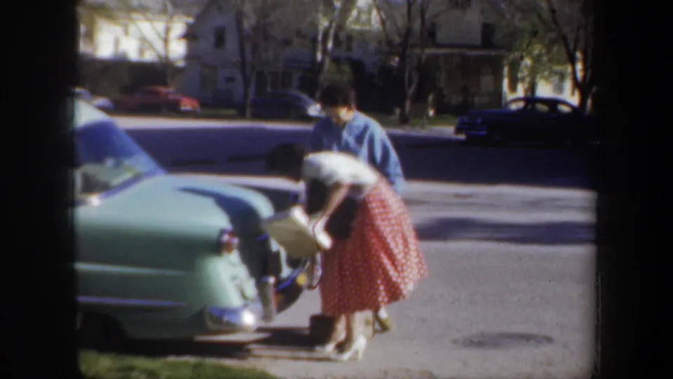 A home-movie frame shows the woman bending into the open trunk of the light green sedan as the man leans his attention toward the loading action.