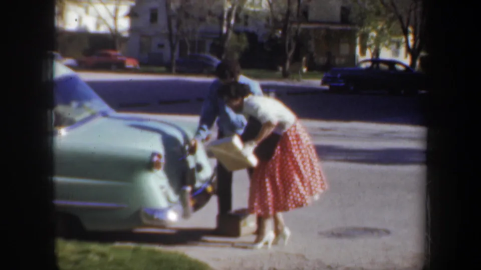 At the end of the short clip, both adults are near the open trunk of the light green sedan, with the box still visible during the loading moment.