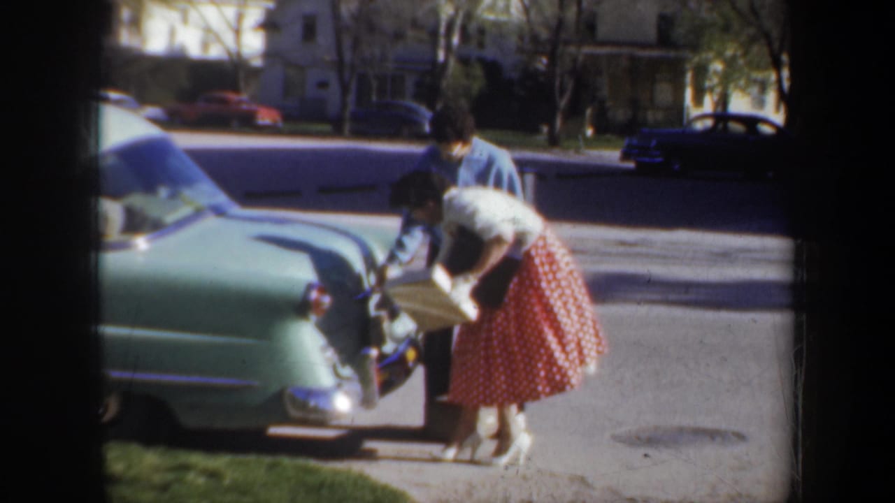 In 1957 Portland home-movie footage, a woman in a blue top and red polka dot skirt bends by a light green car as another person assists.