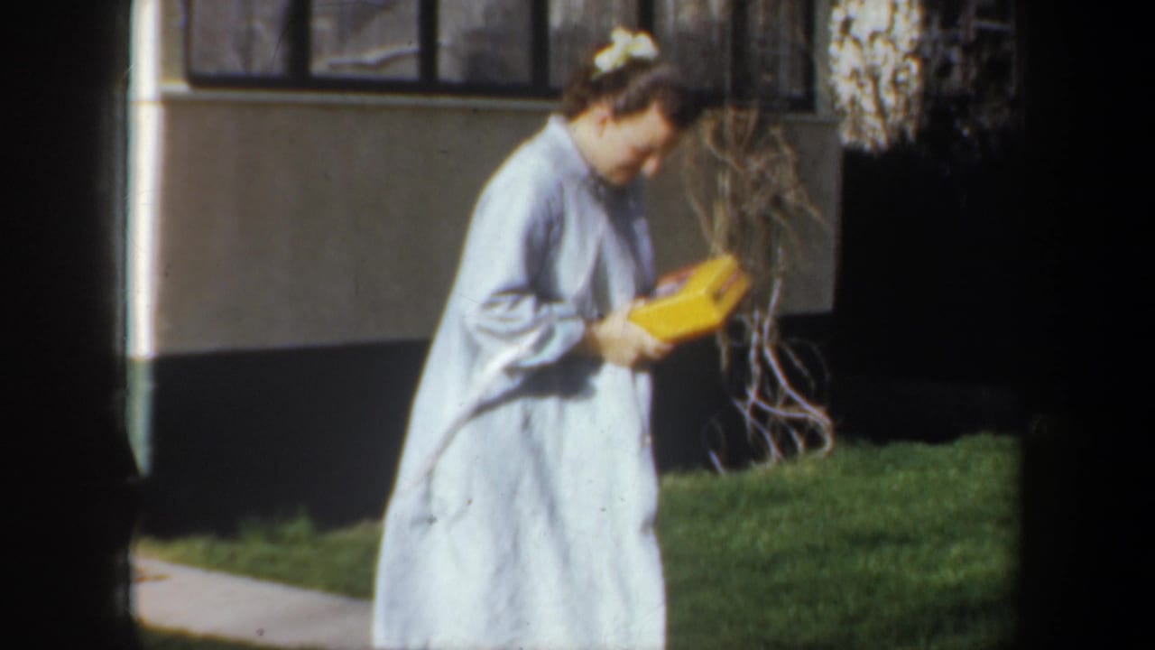 Woman in a pale blue robe carries a yellow box beside a house and lawn.