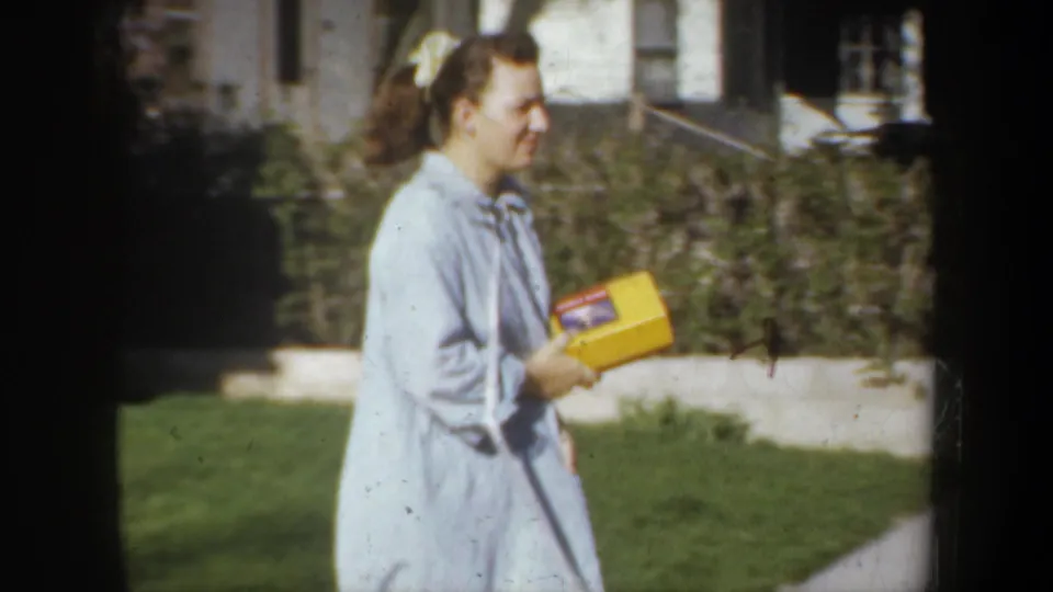 A woman in a pale blue robe holds a yellow box in a 1957 Portland residential yard, with hedge, lawn, and house details behind her.