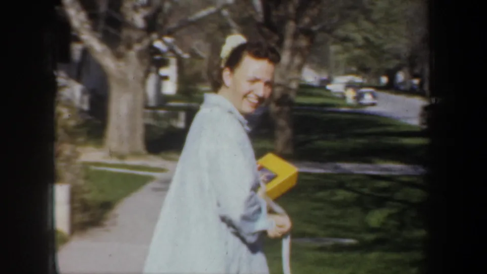 A smiling woman in a pale blue robe carries a yellow box along a Portland sidewalk in 1957 home-movie footage.