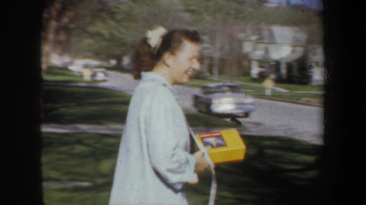 In supplied 1957 Portland footage, a smiling woman with a hair bow carries a yellow box beside lawns and parked cars.