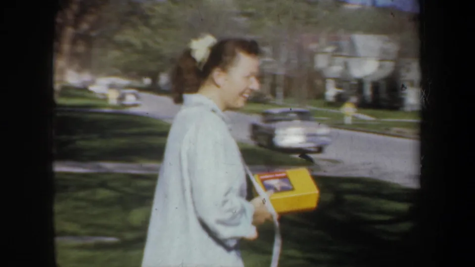 In supplied 1957 Portland footage, a smiling woman with a hair bow carries a yellow box beside lawns and parked cars.