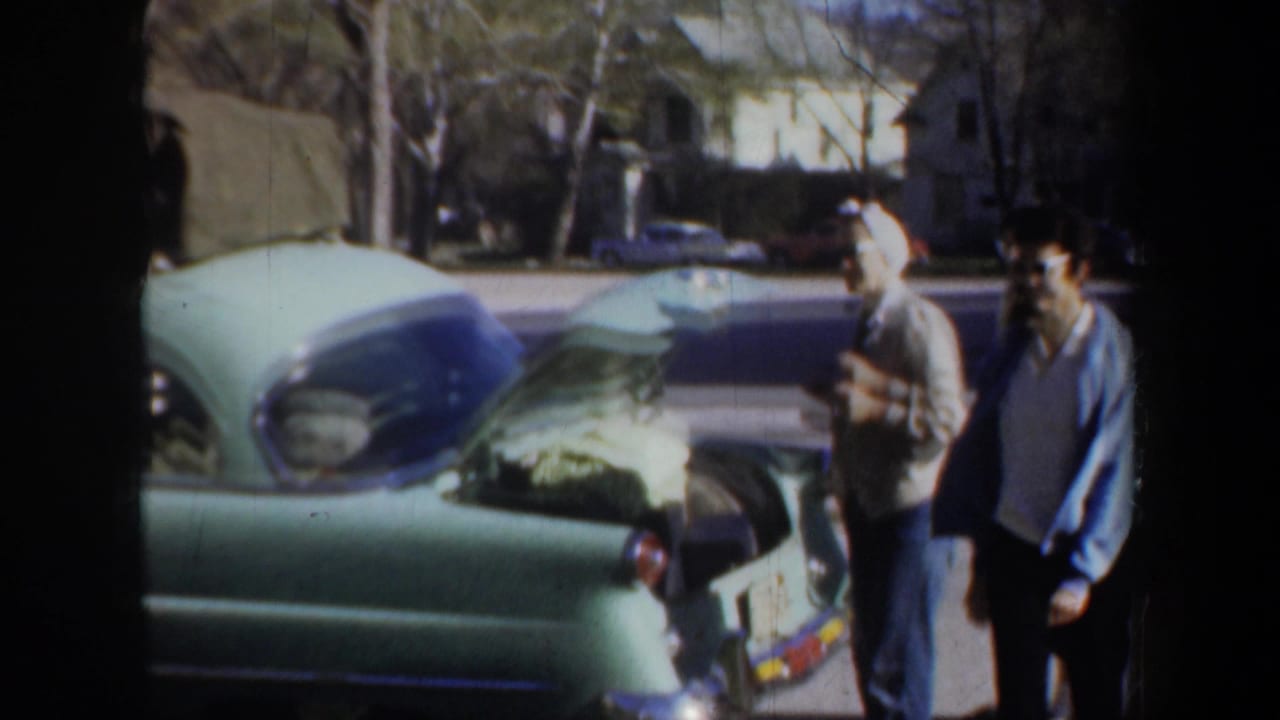 People standing near a light green car with its hood open on a residential street.