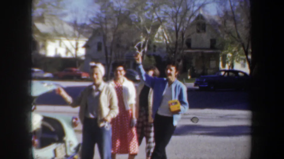 Adults laugh and gesture beside a light green car with its hood open in the supplied 1957 Portland home-movie scene.