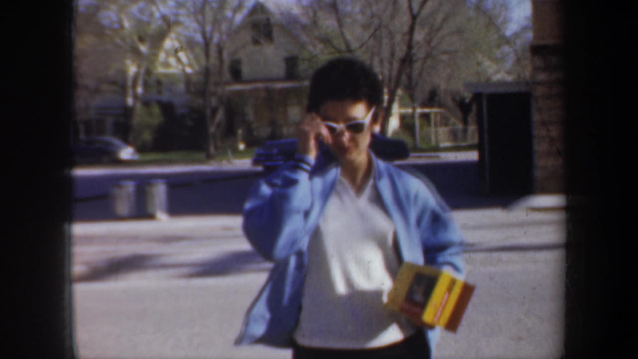 Woman in blue cardigan and white sunglasses walks outdoors holding a yellow box.