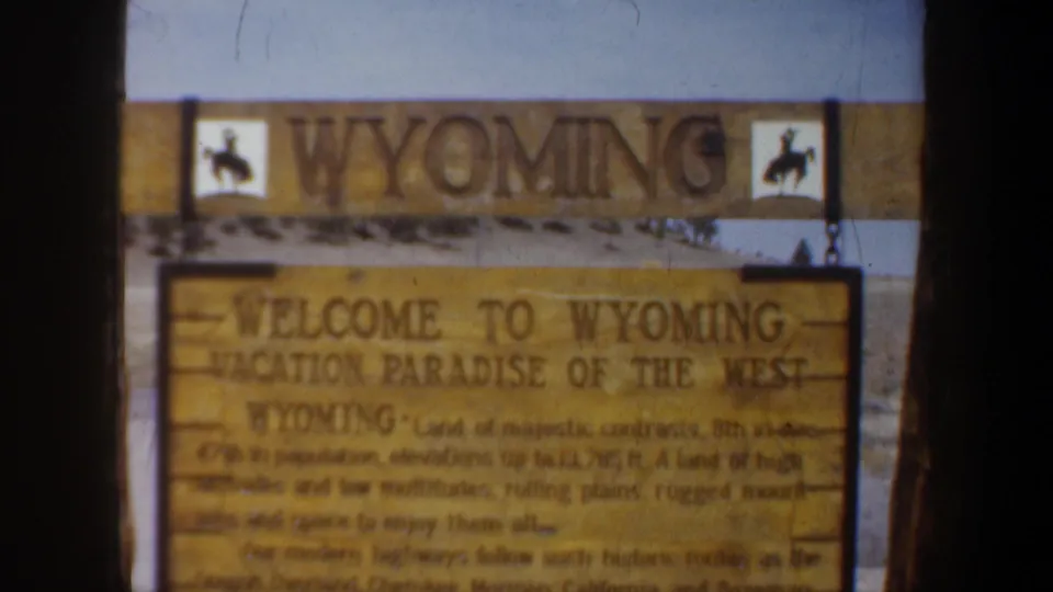 A passing vehicle view holds on the Wyoming welcome sign with cowboy icons and a broad dark divider in the foreground.