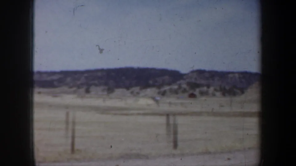 The view shifts from the sign to open prairie with fence posts and low hills beyond the vehicle window.