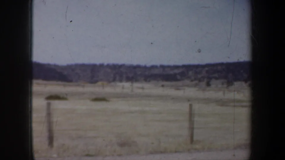 A soft 8mm passenger view of prairie fields, a simple fence line, and distant low hills under a pale sky.