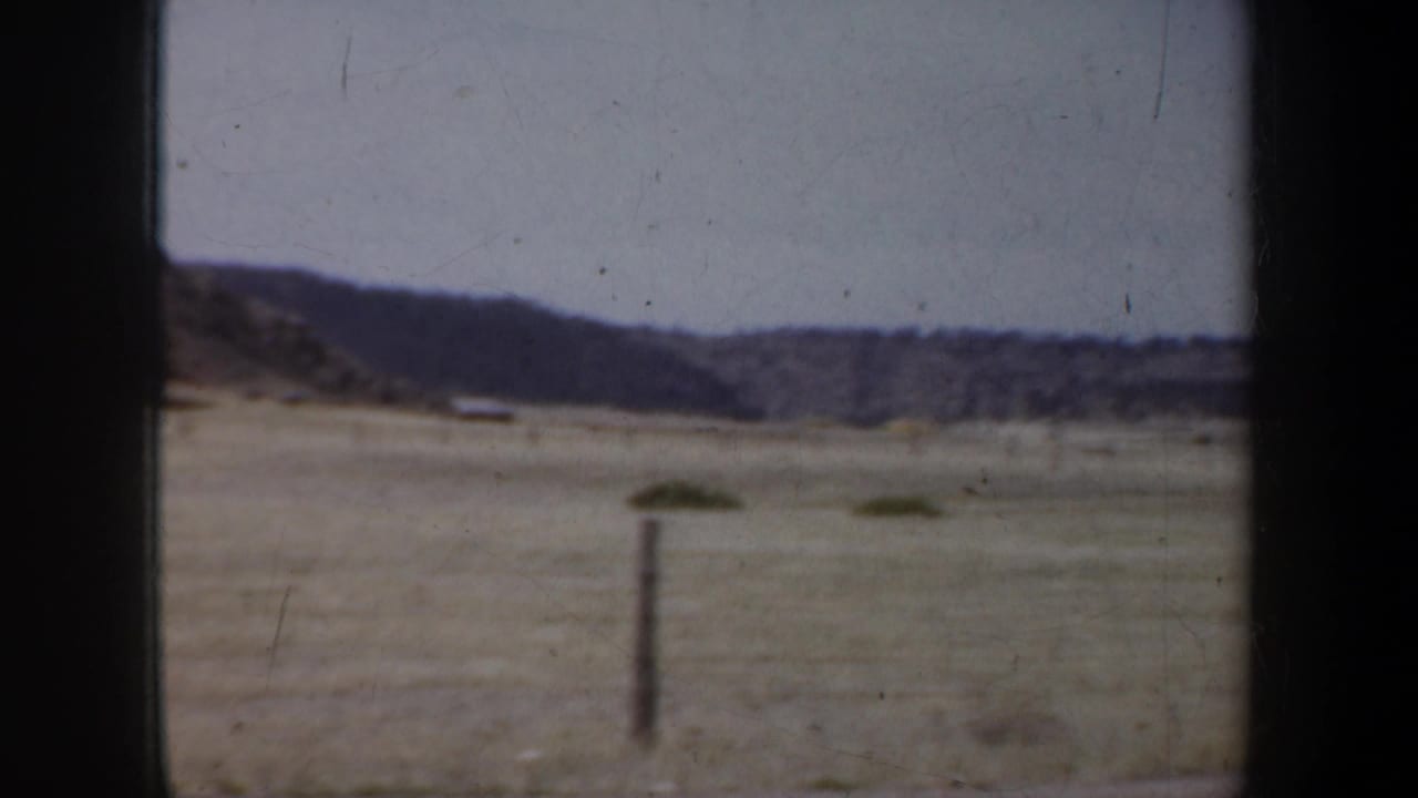 Prairie grass and distant hills seen through a dark vehicle window frame.