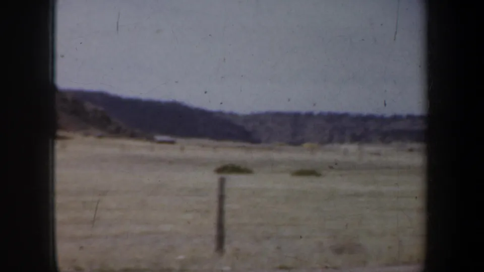 1957 8mm home-movie frame of open prairie, a fence post, and low hills viewed through a dark car-window edge near the reported Portland, Oregon travel sequence.