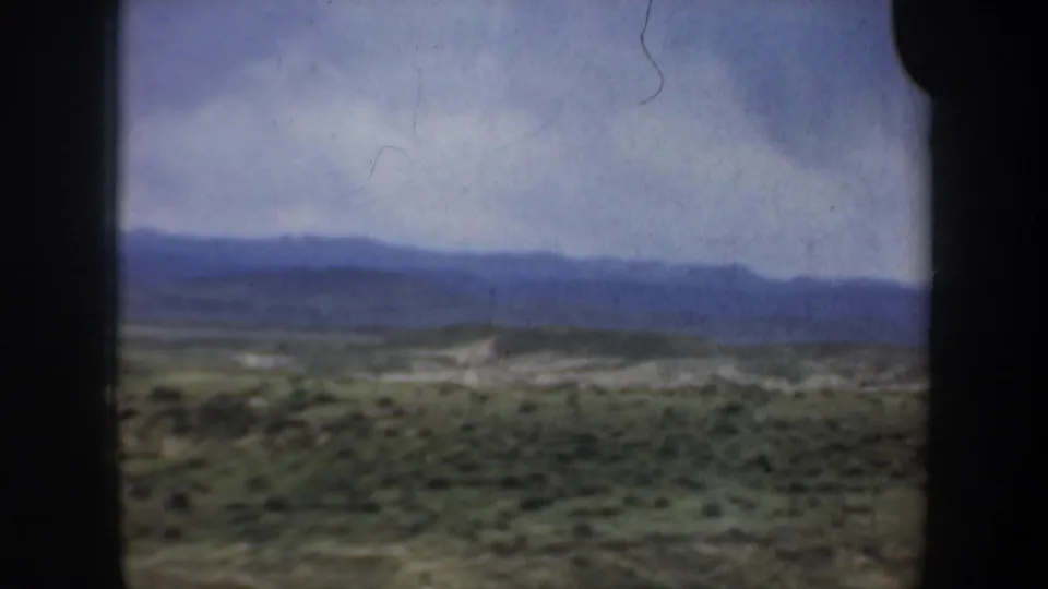 1957 8mm travel view of distant blue ridgelines, open scrub-covered ground, and a pale cloudy sky seen from a moving vehicle.