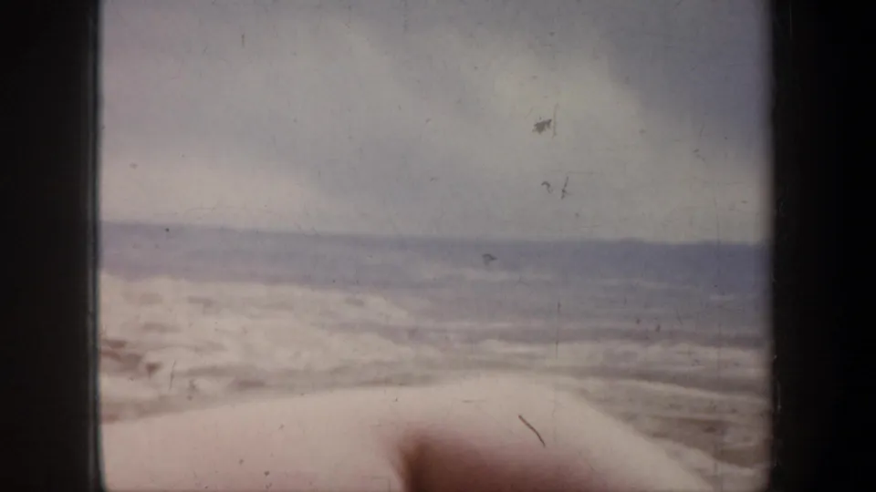1957 8mm home-movie frame with a soft, passing view of open land and low hills under gray cloud cover.