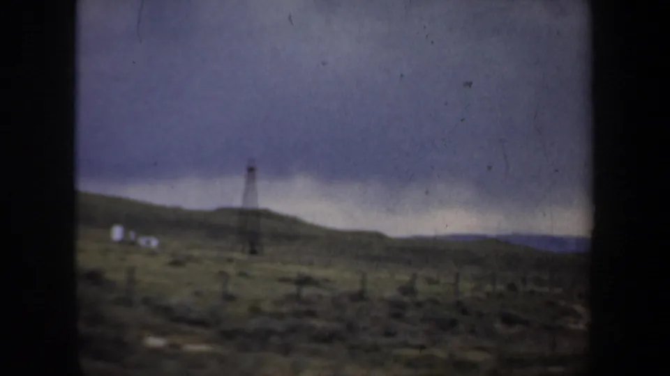 A 1957 Oregon home-movie view appears to pass a distant utility or oil-field structure on a low hillside beneath heavy clouds.