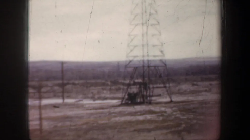 A tall lattice transmission tower and roadside utility poles appear in a soft 1957 8mm view of open industrial land.