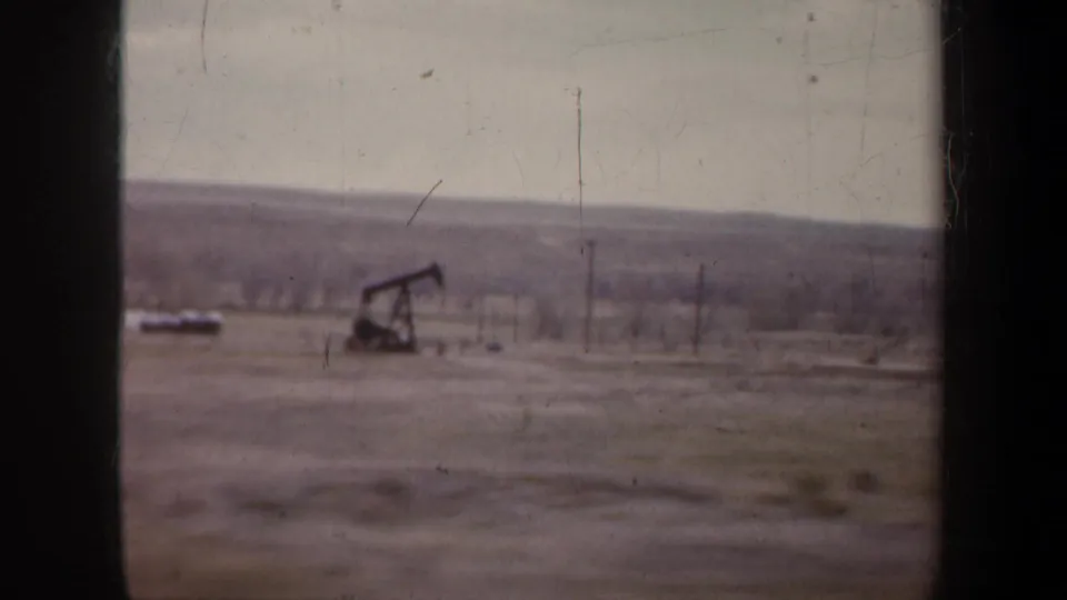 A distant oil pump jack appears in a 1957 home-movie frame, set in open land with utility poles and low hills under a pale sky.