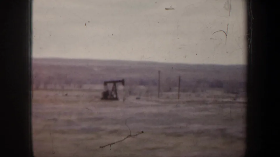 1957 8mm home-movie frame of a rural field with an oil pump jack and utility poles, viewed through dark window framing.
